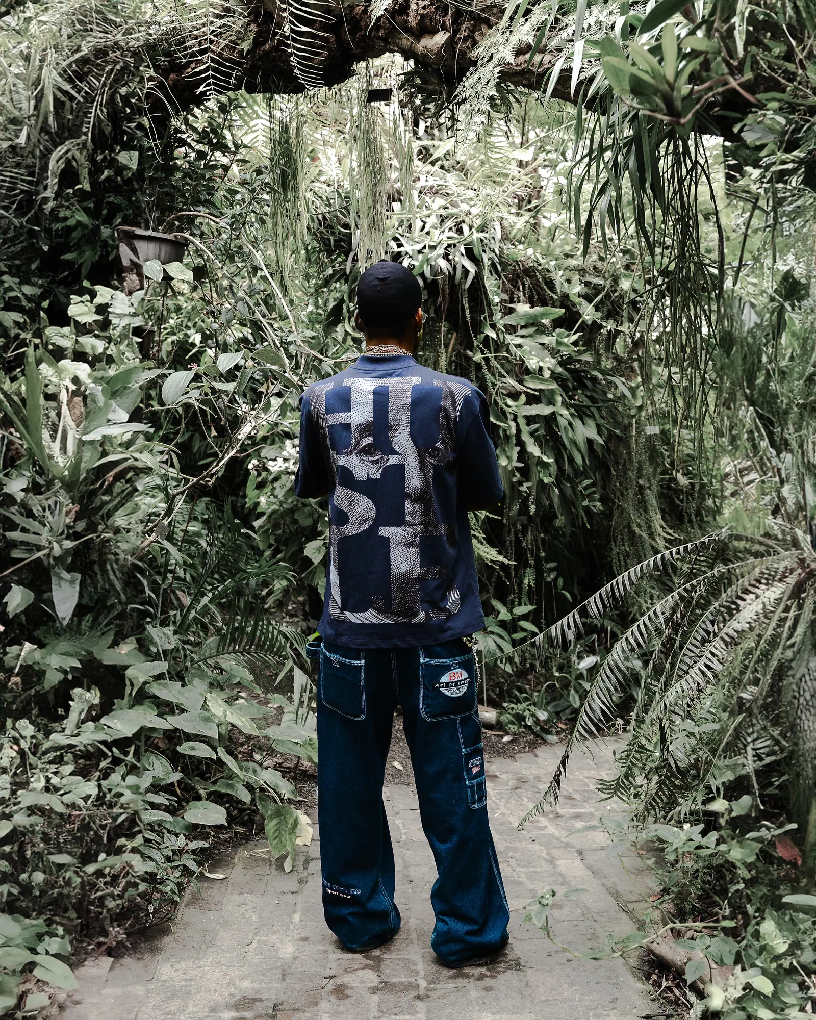 A man seen from behind on an overgrown greenhouse path, in a navy T-shirt with a HUSTLE back print and wide jeans.