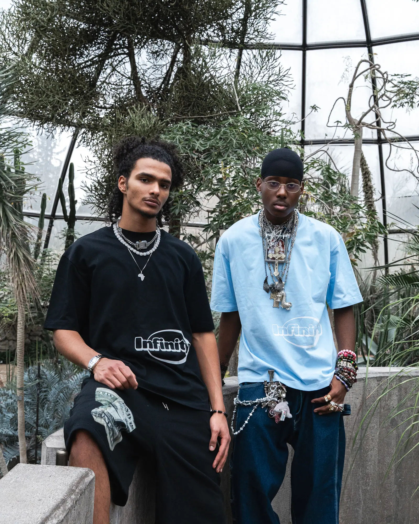 Two men sit on a low wall inside a glasshouse, one in a black Infinityy T-shirt, the other in light blue, both wearing layered chains.