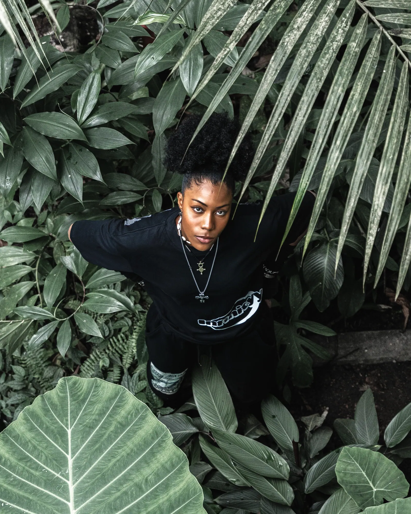 A woman in a black Infinityy T-shirt looks up at the camera from between large tropical leaves.