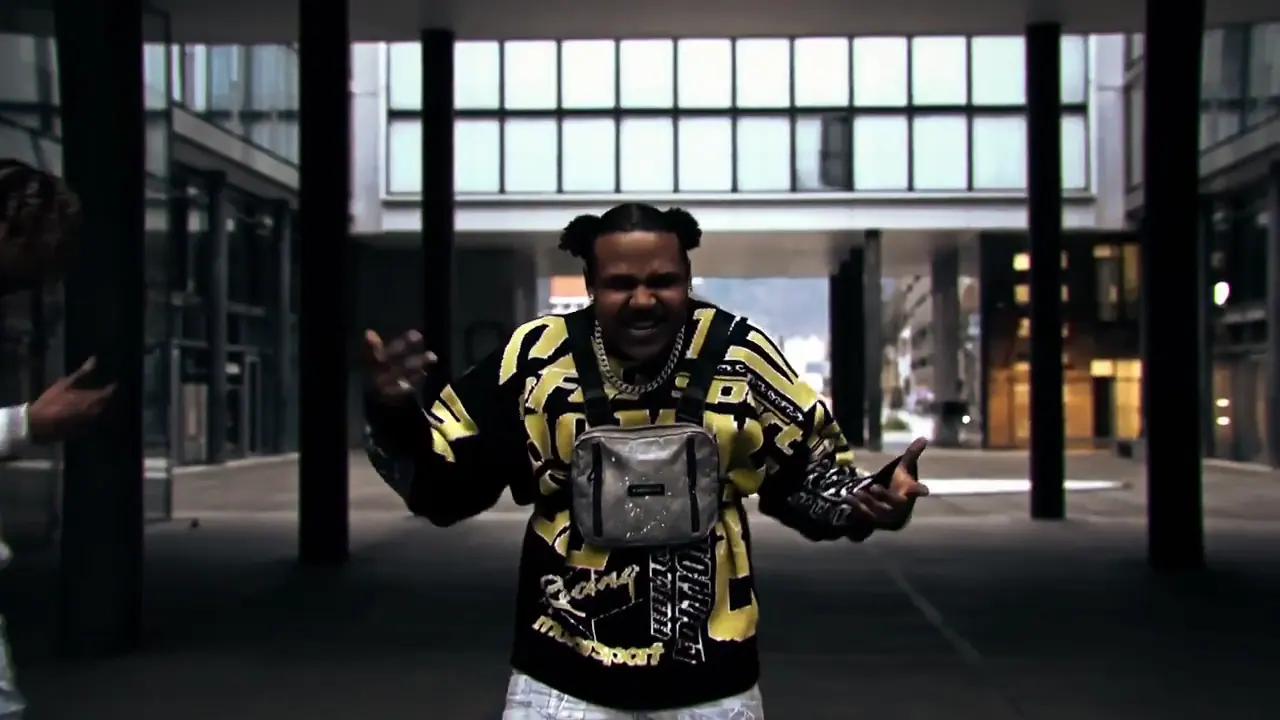 Still from the music video: a rapper in a black-and-yellow patterned jacket with a chest bag stands with his arms open in a street lined with columns, glass buildings behind him.