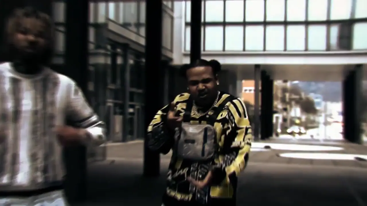 Still from the music video: a rapper in a black-and-yellow jacket with a chest bag walks toward the camera under a concrete overpass, a man in a patterned knit sweater in the foreground.