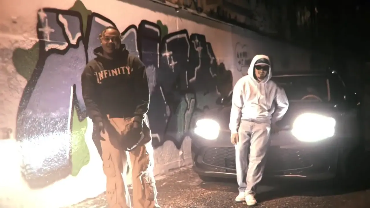 Still from the music video: at night in front of a graffiti wall, a man in a dark Infinityy hoodie and a figure in a white hoodie and sunglasses stand before a car with its headlights on.