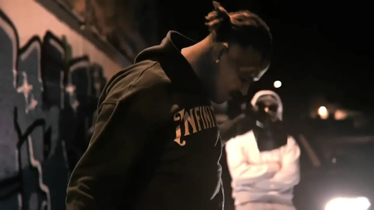 Still from the music video: a man in a dark Infinityy hoodie bows his head in front of a graffiti wall at night, a figure in a white hoodie behind him in the headlights.