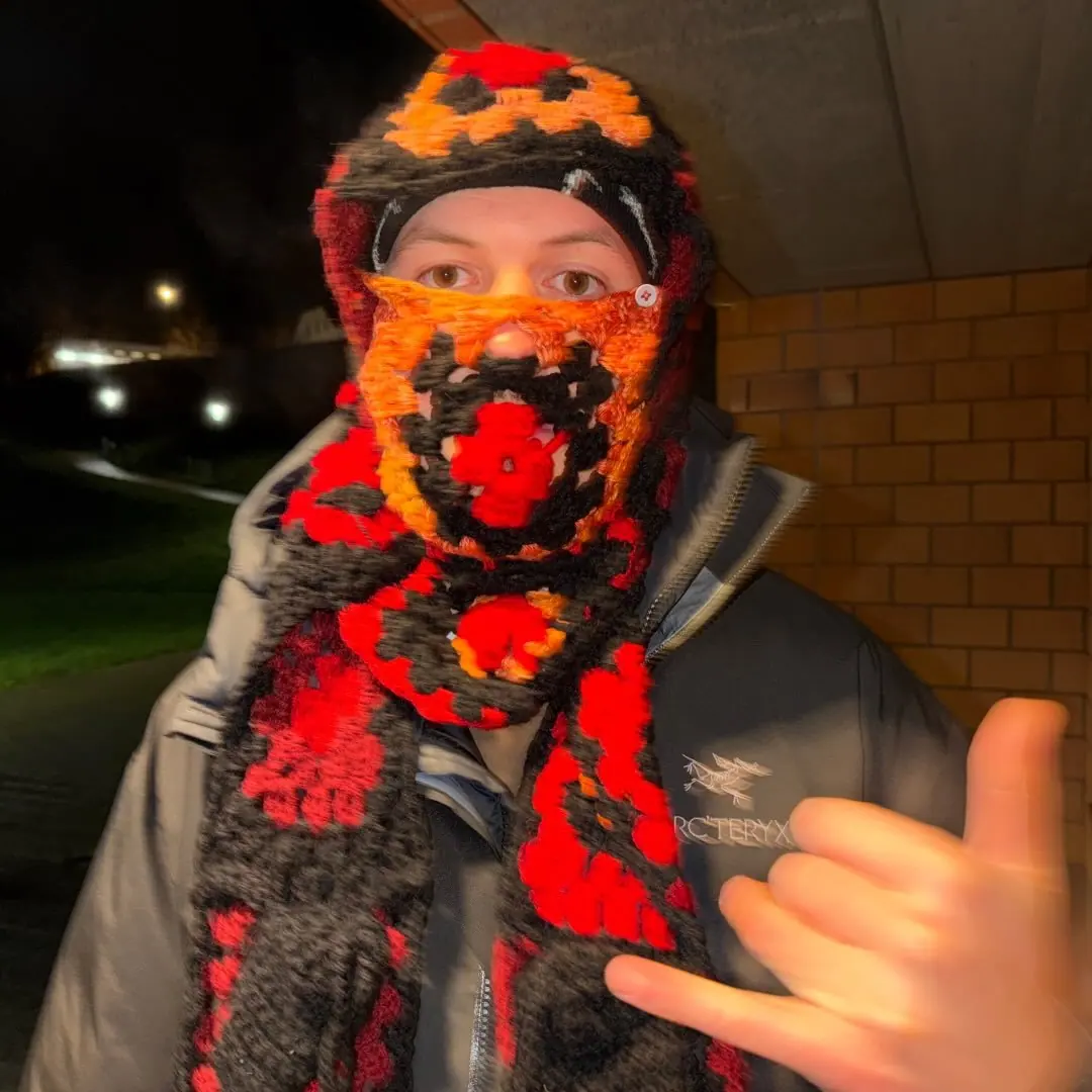 Nachts unter einem Backsteinvorsprung: Eine Person mit gehäkelter rot-orange-schwarzer Sturmhaube und passendem Schal blickt in die Kamera und macht ein Shaka-Zeichen.