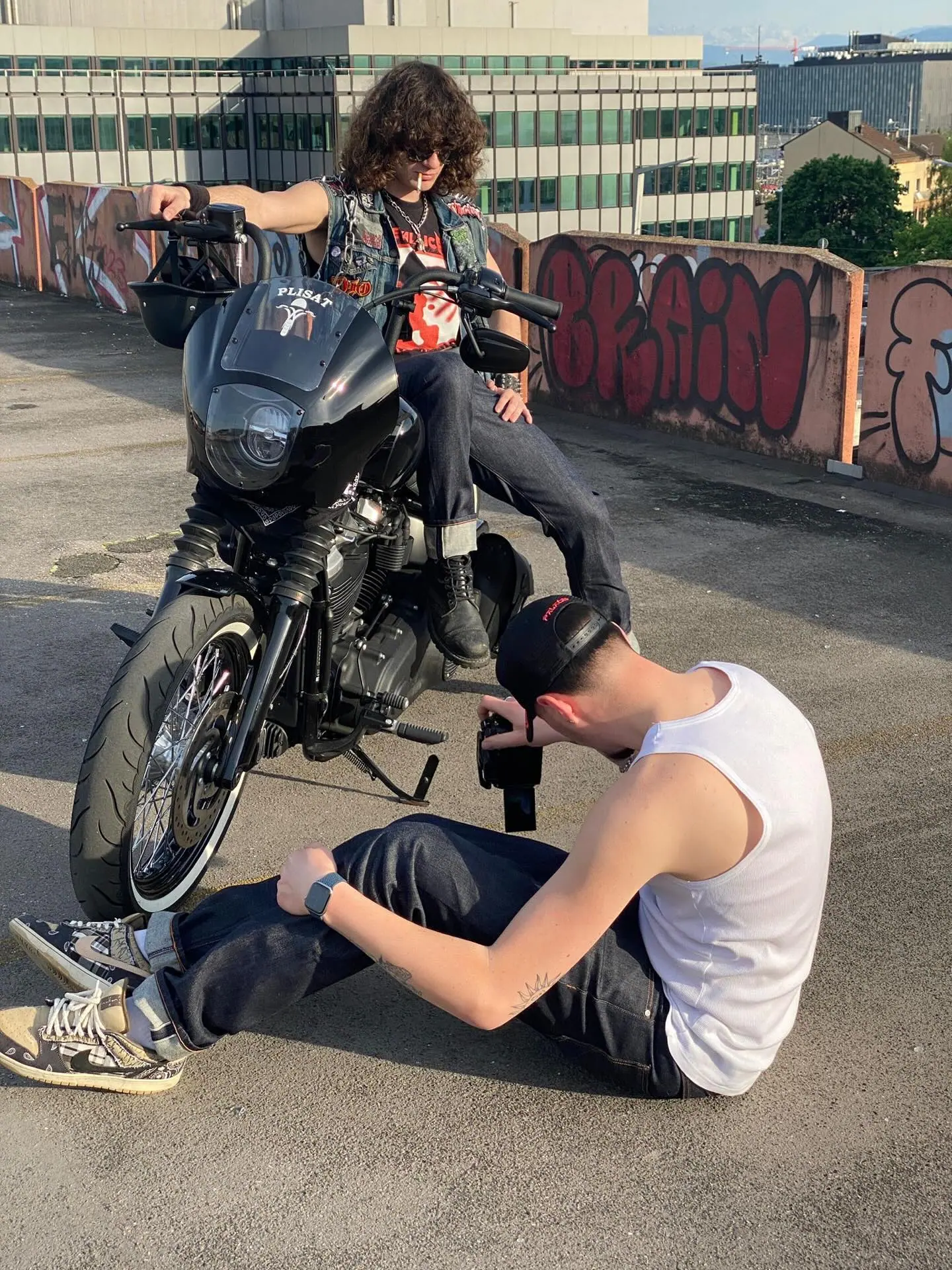 Hinter den Kulissen auf einem Parkdeck: Ein Fotograf in weissem Tanktop und schwarzer Cap sitzt am Boden und fotografiert von unten einen langhaarigen Fahrer mit Sonnenbrille und einer Jeansweste mit Aufnähern auf einem schwarzen Motorrad, dahinter mit Graffiti bedeckte Wände und Bürogebäude.