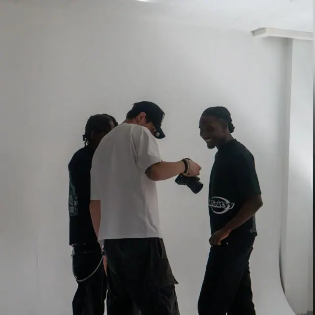 Martin, in weissem T-Shirt und schwarzer Cap, prüft in einem weissen Fotostudio ein Bild auf seiner Kamera, neben ihm zwei Personen in schwarzen T-Shirts.